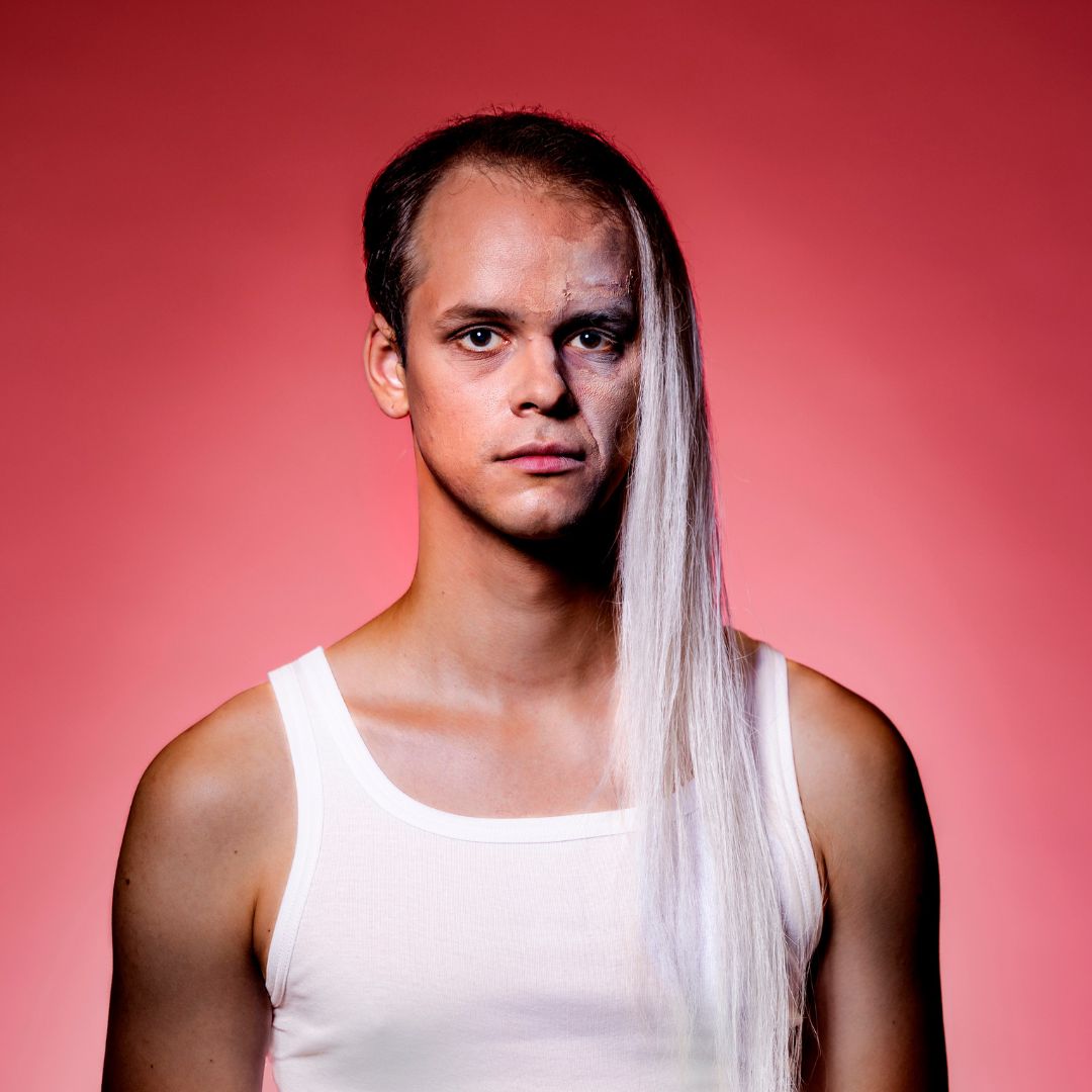Plakatmotiv Herkunft, Foto: Jochen Quast Kolja Schumann mit langem, silberweißem Haar und weißem Tanktop vor rosafarbenem Hintergrund. Foto: Jochen Quast