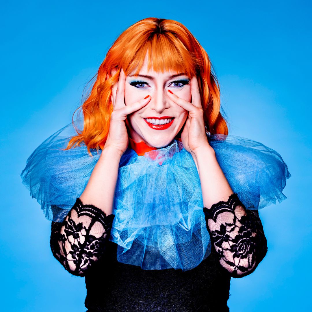 Plakatmotiv Die Fledermaus, Foto: Jochen Quast Sarah Hanikel mit leuchtend orangefarbenem Haar und rotem Lippenstift in blauem Tüllkostüm, die mit den Händen ihr Gesicht umrahmt., Foto: Jochen Quast