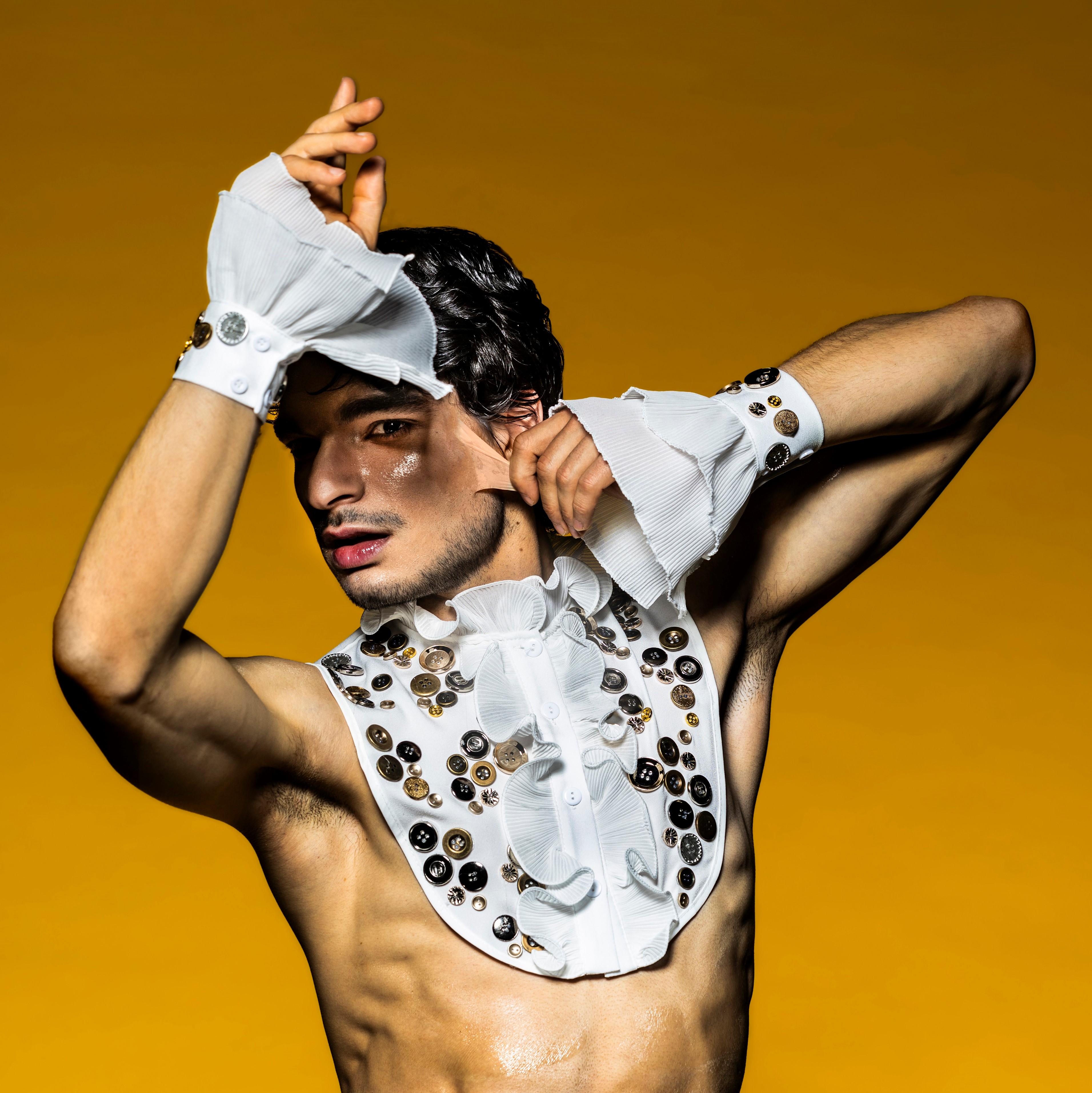 Plakatmotiv Peer Gynt, Foto: Jochen Quast Junger Mann (Pau Pérez Piqué) trägt ein mit Knöpfen verziertem Rüschen-Outfit vor gelbem Hintergrund. Im Gesicht reißt er sich die Haut von der Wange, Foto: Jochen Quast
