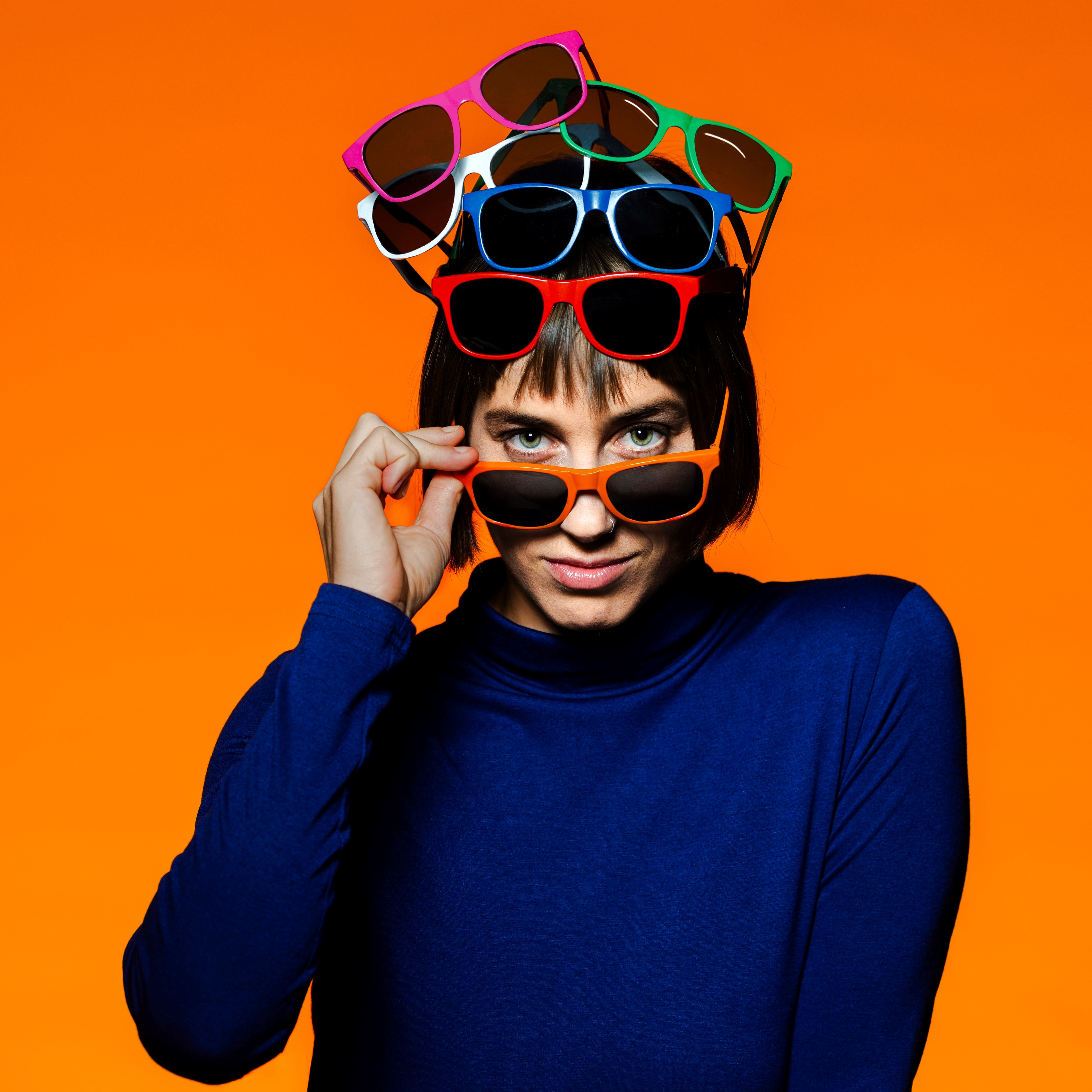 Plakatmotiv Vor Sonnenaufgang, Foto: Jochen Quast Eine Frau (Hannah Rang) mit Sonnenbrille und blauem Pulli schaut in die Kamera. Auf ihrem Kopf stapeln sich weitere Sonnenbrillen., Foto: Jochen Quast